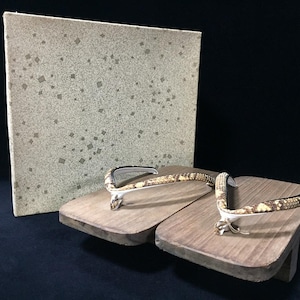 May include: A pair of wooden geta sandals featuring a natural wood grain base and thong straps with a textured snakeskin pattern. The sandals are displayed in front of a rectangular box with a light beige surface and small square gold accents.
