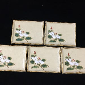 May include: Five rectangular ceramic plates with a beige background and a floral design. Each plate features a white flower with green leaves and a red bud. The edges are trimmed with a brown border.