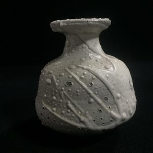 May include: A small, off-white ceramic vase with a textured surface and a bulbous body. The vase has a narrow neck and a flared rim. The surface is decorated with raised dots and diagonal lines, creating a unique, handcrafted appearance.