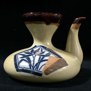 Puede incluir: Una botella de sake de cerámica con una forma única y un pico. La botella es beige con un borde y punta de pico marrón. Presenta un diseño floral azul y blanco y un patrón geométrico marrón y beige.