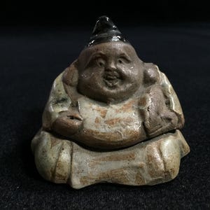 May include: A small, ceramic figurine of a seated, smiling figure with a black hat. The figure has a round body and is wearing a light-colored robe. The figurine is primarily brown and beige with some darker accents.