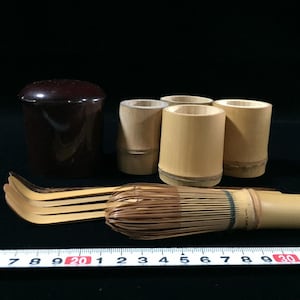 May include: A collection of tea ceremony tools including a dark brown lidded container, three cylindrical bamboo cups, a bamboo whisk, and three curved bamboo scoops. A white measuring tape is visible in the foreground, showing a scale in centimeters.