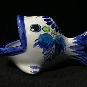 May include: A white ceramic fish-shaped bowl with a wide open mouth. The exterior features hand-painted floral patterns in shades of blue and green, with a solid blue rim around the mouth and a blue-tipped tail fin on the right side.