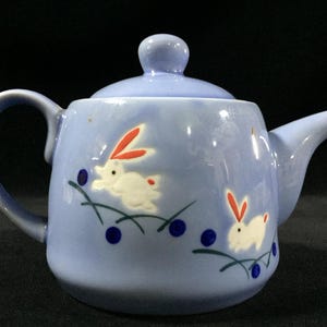 May include: A pale blue ceramic teapot with a lid and handle. The teapot features white rabbits with red ears and blue berries. A delightful item for serving tea, adding a touch of whimsy.