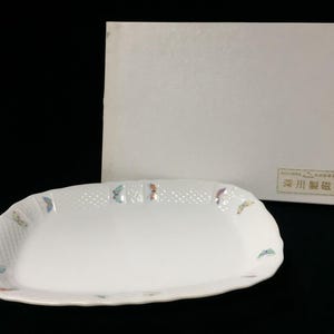 以下が含まれることがあります： 角が丸い長方形の白い磁器製サービングプレート。縁には格子状のテクスチャが施され、小さなカラフルな蝶のイラストが描かれています。深川製磁と書かれた白い収納箱が付属しています。