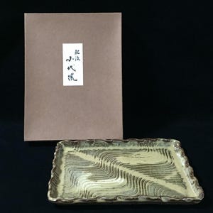 May include: A rectangular ceramic tray with a wavy, cream and dark brown linear pattern and a textured, uneven rim. Behind the tray stands a rectangular tan cardboard box featuring a white label with black Japanese calligraphy characters.