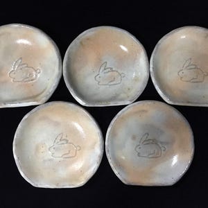 Peut inclure: Cinq petits plats en céramique ronds avec un vernis brillant aux tons blanc cassé et beige marbré. Chaque plat présente au centre un dessin au trait simple et gravé représentant un lapin assis. Les bords sont légèrement irréguliers.