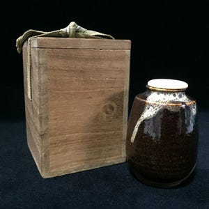Pode incluir: Um pequeno pote de cerâmica castanho-escuro com acabamento brilhante e um padrão de esmalte branco escorrido à volta da borda, acompanhado por uma caixa de arrumação quadrada de madeira clara com um fecho de tecido no topo.