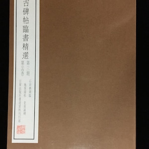 May include: A brown rectangular book with a vertical white label on the left side. The label features black Chinese characters reading 古碑帖臨書精選, 第三期, 第三十四卷, 石田栖湖臨, 瘞鶴銘, 皇甫誕碑, and 日貿出版社書道資料刊行部, with a small red square seal at the bottom.