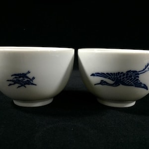 May include: Two white ceramic bowls with a blue crane design. One bowl features two cranes in flight, the other a single crane. The bowls are set against a dark background.