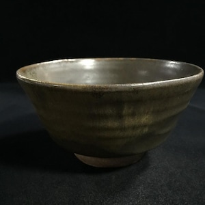 May include: A handmade ceramic bowl with a textured, ridged exterior in shades of olive green and brown. The interior has a lighter, almost metallic, gray hue. The bowl has a rounded shape and sits on a small, circular base.