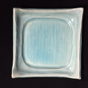 以下が含まれることがあります： 光沢のあるライトブルーの釉薬が施された正方形の陶器皿。わずかに盛り上がった波状の縁と、滑らかで凹状の中央部が特徴です。中央部には繊細な縦のくぼみがあり、皿の表面全体に質感のある外観を作り出しています。
