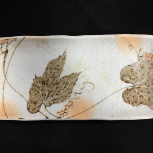 May include: Rectangular ceramic serving dish with a white background and a brown leaf and vine design. The dish has a slightly textured surface and rounded edges. Peach-colored accents are also present.