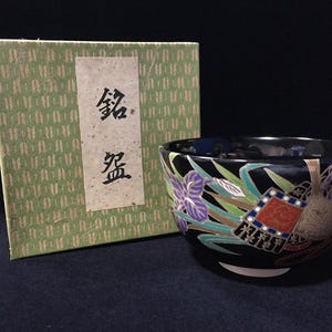 以下が含まれることがあります： 黒いセラミックボウル、花柄と幾何学模様。緑色の箱には日本語のテキストが書かれています。ボウルには紫色のアイリス、緑の葉、赤と青の幾何学模様があります。箱には繰り返し模様があります。