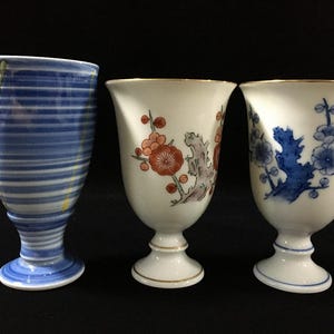 以下が含まれることがあります： 脚付きの陶器製カップ3点。左のカップは青と白の横縞模様に黄色の縦のアクセントが入っています。中央のカップは白地にオレンジとグレーの花枝模様が描かれています。右のカップは白地に青い花枝模様が描かれています。