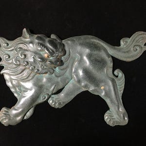 May include: A dark bronze-toned metal wall plaque depicting a stylised guardian lion in a prowling stance. The figure features intricate swirling mane details, a long flowing tail, and a weathered patina finish with subtle hints of green oxidation.