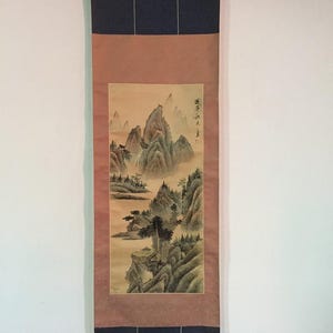 May include: A vertical hanging scroll featuring a landscape painting of jagged mountains, pine trees, and mist. The artwork is framed by a tan border and dark blue fabric panels at the top and bottom, with a black wooden rod at the base for hanging.