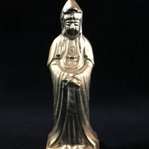 May include: A gold-colored statue of a robed figure with hands clasped in front. The statue has a serene expression and is standing on a rectangular base. The figure is wearing a hooded garment.