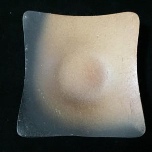 Op de afbeelding: Een vierkante keramische schaal met een concaaf midden, voorzien van een kleurverloop van diep houtskoolzwart aan de ene kant naar een warm, zandkleurig beige aan de andere kant, met een getextureerde, matte afwerking over het gehele oppervlak.