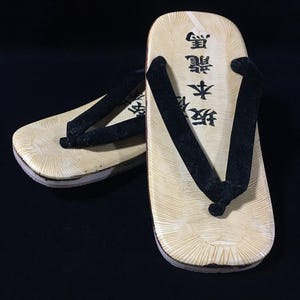 May include: A pair of geta sandals featuring light beige wooden footbeds with a radial grain pattern and black fabric thong straps. The footbeds are printed with black Japanese characters reading 坂本龍馬. The sandals have a layered base construction.