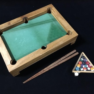 以下が含まれることがあります： 緑色のフェルト面、4つのコーナーポケット、2本の木製キューを備えた、ミニチュア木製ビリヤード台。木製の三角形には、カラフルなビリヤードボールのセットが入っています。あらゆる年齢層向けの楽しいゲームです。