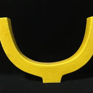 May include: A bright yellow, U-shaped decorative object with a smooth, matte finish. The piece features a wide, curved design that tapers slightly toward the top ends and rests on a small, rectangular base centered at the bottom of the curve.