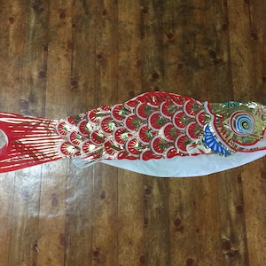 May include: A red, gold, and white carp-shaped windsock featuring a detailed scale pattern across the body, a prominent eye design near the circular opening, and a long, flowing tail section with red stripes, designed for outdoor display.