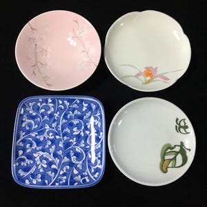 Può includere: Un set di quattro piatti in ceramica composto da una ciotola rotonda rosa chiaro con motivi floreali bianchi, una ciotola rotonda bianca con un fiore rosa e arancione, un piatto quadrato blu e bianco con volute e un piatto rotondo bianco con foglie verdi.