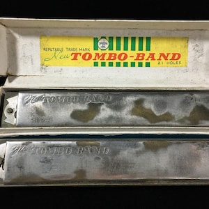 Pode incluir: Duas harmônicas de metal prateado rotuladas como New Tombo-Band 21 Holes, apresentadas em uma caixa de papelão branca. A tampa da caixa apresenta um rótulo amarelo e verde com o texto Reputable Trade Mark New Tombo-Band 21 Holes e um logotipo circular.