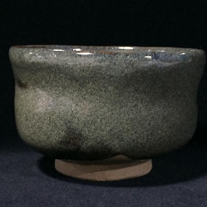 May include: A ceramic bowl with a speckled, mottled green and grey glaze. The bowl features a rounded, slightly irregular shape with a thick rim and a distinct, unglazed circular base that is light tan in color, contrasting with the dark body.