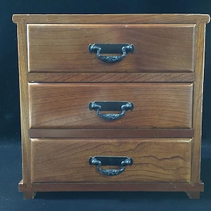 Könnte beinhalten: Eine kleine Holzkommode mit drei horizontalen Schubladen, die jeweils mit einem dunklen Metallgriff mit verzierten Details versehen sind. Das Holz hat eine warme braune Oberfläche mit sichtbarer Maserung und steht auf vier kurzen Füßen.