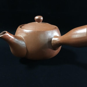 May include: A brown clay teapot with a smooth, matte finish. The teapot features a rounded body, a short spout, a lid with a small knob handle, and a prominent side handle that extends outwards, designed for a comfortable grip while pouring tea.