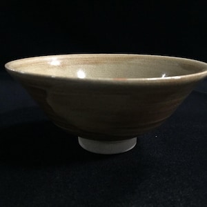 May include: A ceramic bowl with a wide, shallow design. The bowl has a light brown interior and a darker brown exterior with visible texture. It sits on a small, white base, and the rim is slightly lighter in color.