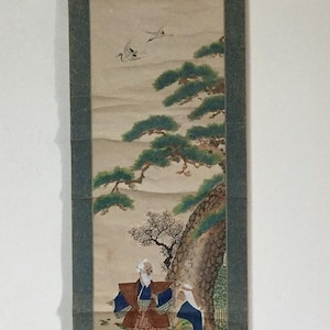 以下が含まれることがあります： 2 人の人物が木の下にいる様子を描いた日本の巻物絵画。空には鶴が飛んでいる。このアートワークは、詳細な筆致と、落ち着いた色合いの穏やかな風景が特徴です。巻物は濃い緑色の縁で縁取られています。