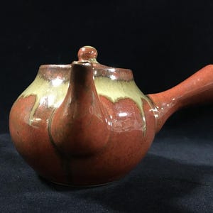 Può includere: Una teiera in ceramica con manico laterale caratterizzata da un corpo arrotondato con una smaltatura lucida color rosso-marrone. La parte superiore della teiera presenta un motivo decorativo a goccia color crema che scende dal bordo.
