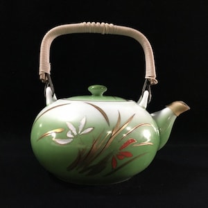 May include: A ceramic teapot featuring a gradient green finish that transitions to white. The body is decorated with painted white flowers, gold-toned grass motifs, and small red accents. It includes a matching lid and a curved, light-coloured woven handle.