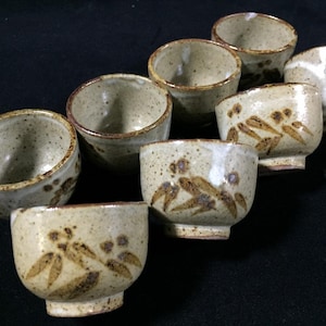 May include: A collection of seven small, handmade ceramic bowls. The bowls are a speckled beige color with brown floral designs and a dark brown rim. The bowls are arranged in a staggered pattern.