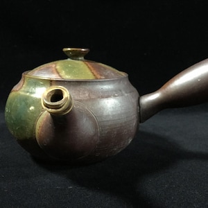May include: A ceramic teapot with a dark brown body, a green and brown lid, and a brown handle. The spout is cylindrical and angled. The teapot has a round shape and a small knob on the lid.