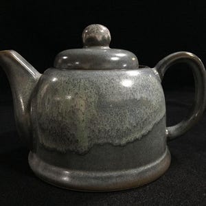 May include: A ceramic teapot featuring a rounded body and a matching lid with a knob handle. The glaze displays a mottled blend of dark charcoal grey and lighter, dusty blue tones, with a smooth, glossy finish and a curved spout and handle.