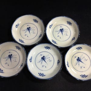 Può includere: Un set di cinque piccoli piatti rotondi in ceramica con base color crema e un motivo a libellula dipinto a mano in blu al centro. Ogni piatto presenta un bordo smerlato e ulteriori segni decorativi blu lungo il bordo interno.
