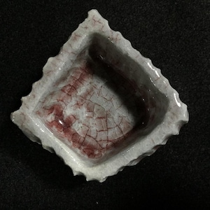 以下が含まれることがあります： 縁が粗くギザギザした四角い陶器の小鉢。内側は白い釉薬で仕上げられており、底面と側面に赤いひび割れ模様がはっきりと入っているため、表面には質感のある古びたような風合いが表現されています。