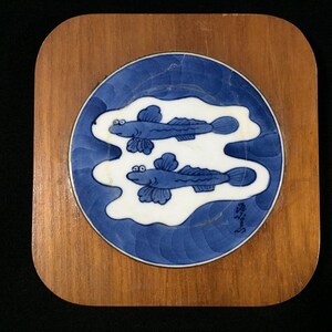 Könnte beinhalten: Ein quadratischer Holzuntersetzer mit abgerundeten Ecken. Er hat einen blau-weißen Keramikeinsatz mit zwei stilisierten blauen Fischen, die in einem weißen Wassermuster schwimmen. Die Fische haben detaillierte Flossen und Augen.