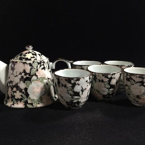Pode incluir: Um conjunto de chá de cerâmica composto por um bule com tampa e quatro chávenas a condizer. Cada peça apresenta um fundo preto decorado com um padrão denso de flores brancas e rosa pálido e folhas verdes, com um rebordo prateado.