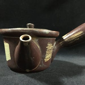 以下が含まれることがあります： 側面に持ち手が付いた茶色の陶器製ティーポット。本体と持ち手には、滑らかなダークブラウンの釉薬の上に、ベージュ色の筆跡のような質感のある装飾模様が施されており、小さな注ぎ口が付いています。蓋も付属しています。