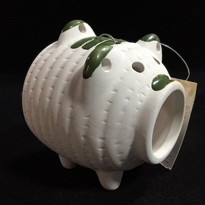 May include: White ceramic pig-shaped planter with a textured surface and green accents. The planter has a large opening for planting and small holes on top. A decorative item for home or garden.