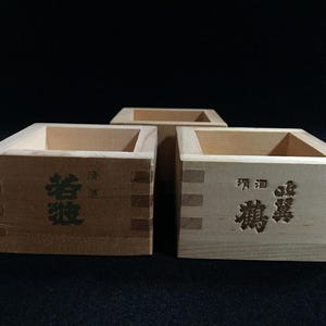 May include: Three square wooden masu cups with light tan grain. The front faces feature dark brown Japanese kanji characters and smaller text. The boxes are constructed with visible interlocking finger joints at the corners and open tops.