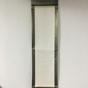 May include: A vertical hanging scroll featuring a blank, rectangular white paper centre panel. The scroll is framed by dark, metallic-toned fabric borders with a repeating intricate pattern, finished with a wooden rod at the bottom edge.