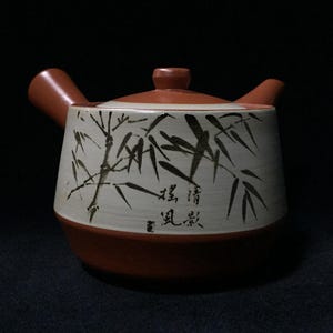May include: A ceramic teapot with a brown base, handle, and lid. The body is white with a bamboo design in black ink and Japanese characters. The teapot has a traditional shape and is likely used for tea.
