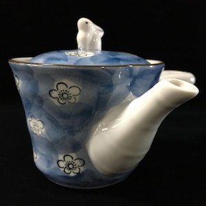 May include: A ceramic teapot with a blue and white watercolor-style floral pattern featuring black outlined blossoms. The lid is topped with a small white rabbit figurine, and the teapot has a side handle and a white spout against a dark background.