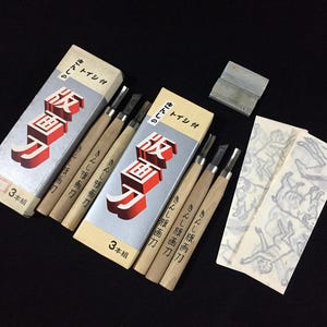 May include: A selection of Japanese wood carving tools, including three boxes of carving knives, a sharpening stone, and printed design templates. The boxes feature red and white Japanese text. The tools are arranged on a black surface.
